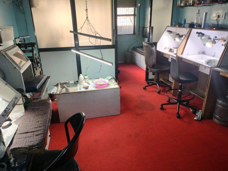 May include: An interior shot of a workshop with specialized equipment. The room features workbenches with built-in lighting, chairs, and a red carpet. A window provides natural light, and various tools and supplies are visible.