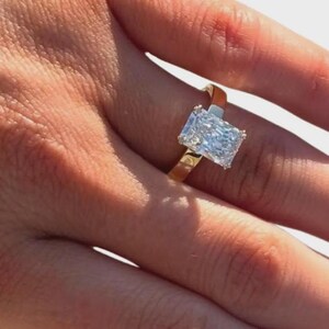 May include: A radiant-cut diamond ring with a gold band. The large, rectangular diamond sparkles, catching the light. The ring is set on a gold band, showcasing a classic and elegant design. A beautiful piece of jewelry.
