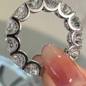 Könnte beinhalten: Ein silberner Eternity-Ring mit einer durchgehenden Reihe klarer, funkelnder Edelsteine. Das Design des Rings zeigt eine Reihe gleichmäßig verteilter, brillantgeschliffener Steine, die einen vollständigen Lichtkreis bilden. Der Ring wird von einer Person gehalten.