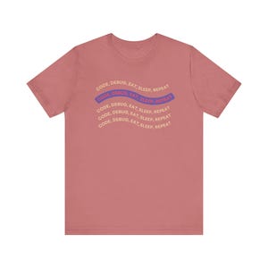 Pode incluir: Camiseta rosa empoeirado com o texto "CODE, DEBUG, EAT, SLEEP, REPEAT" em um design curvo repetido. O texto é multicolorido, com azul, roxo e bege. A camiseta tem mangas curtas e gola redonda.