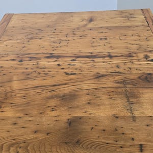 May include: A close-up view of a rectangular wooden table. The table has a natural wood grain with dark knots and imperfections. The edges are framed with a darker wood border. The table is made of solid wood.