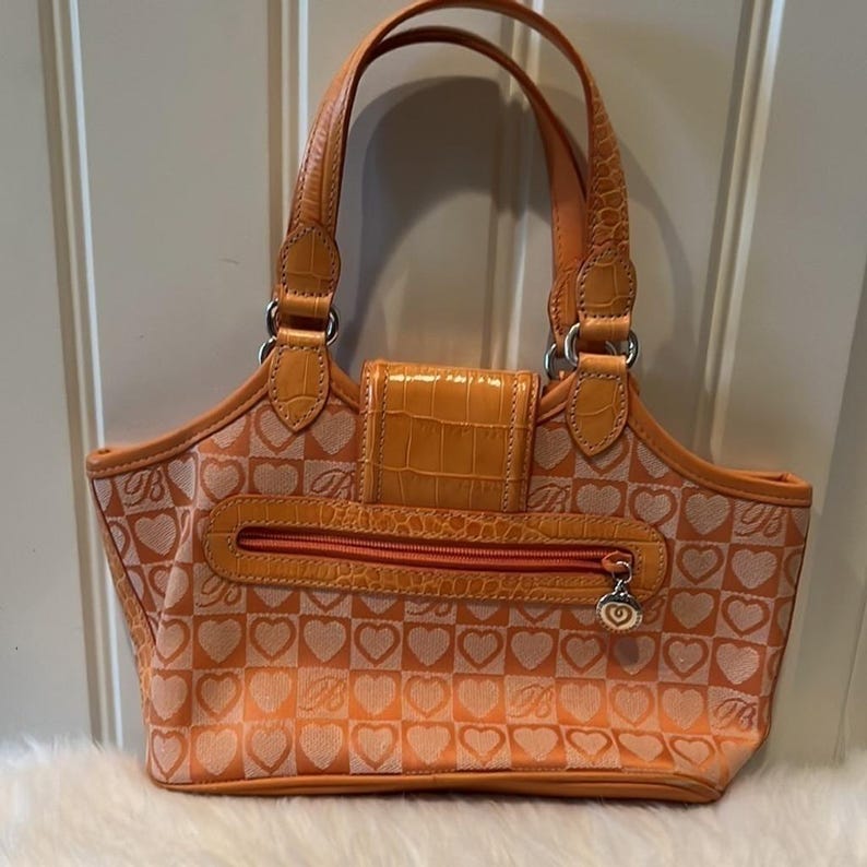 May include: Orange handbag with a heart pattern and a top handle. The bag features a flap closure and a zipper pocket. The bag has a textured leather-like appearance and silver-tone hardware. The bag has a decorative charm on the zipper.
