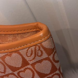 May include: Close-up of a section of a handbag. The bag features a repeating heart pattern in orange and white. The top edge is trimmed with orange leather and stitched with a contrasting thread.