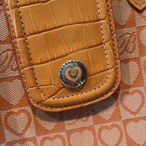 May include: Close-up of a tan leather handbag with a crocodile texture. The bag features a silver heart-shaped emblem and is set against a peach-colored background with a heart pattern. The stitching is visible around the edges of the leather.