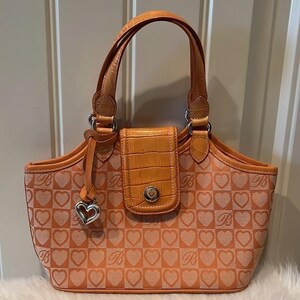 May include: Orange handbag with a heart pattern and silver hardware. The bag features a top handle, a flap closure, and a heart-shaped charm. The bag's exterior is patterned with hearts and the letter "B".