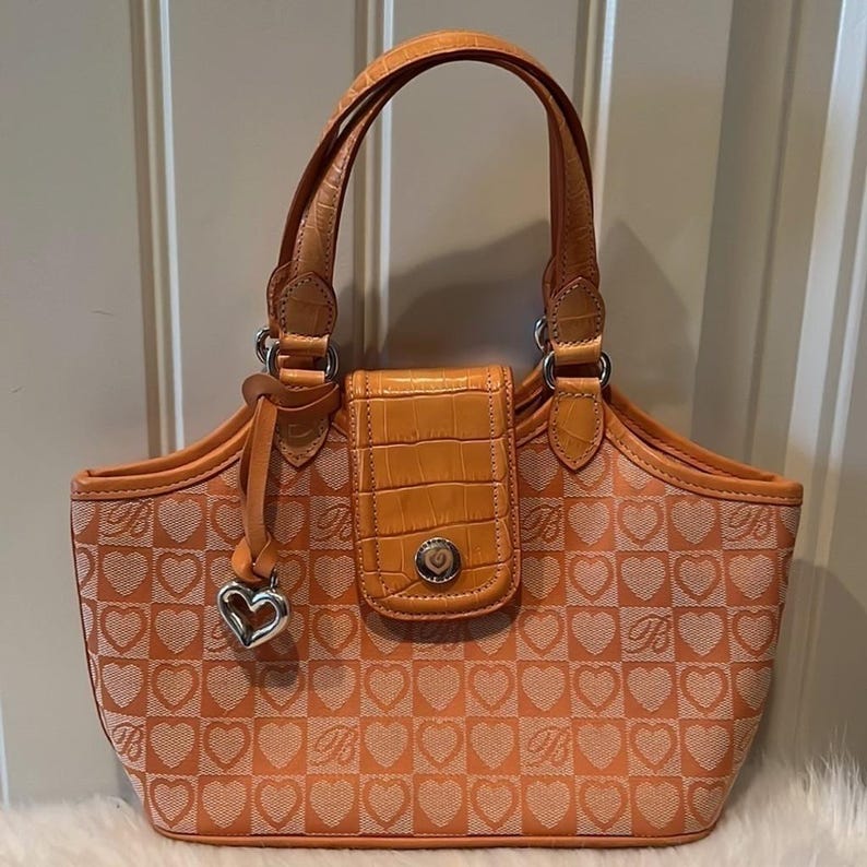 May include: Orange handbag with a heart pattern and silver hardware. The bag features a top handle, a flap closure, and a heart-shaped charm. The bag's exterior is patterned with hearts and the letter "B".
