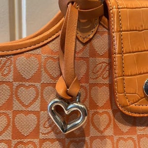May include: Close-up of an orange handbag with a heart pattern. The bag features a silver heart-shaped charm and a textured, crocodile-like leather pocket. The bag's design includes a repeating heart pattern on a checkered background.