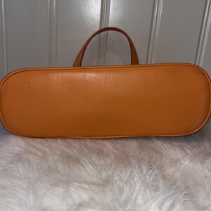 May include: An orange handbag with a rounded rectangular shape and a single handle. The bag is made of a smooth material and sits on a white, fluffy surface. The handle is a lighter shade of brown.