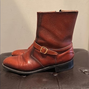 May include: A pair of brown leather ankle boots with a buckle detail. The boots have a rounded toe and a short heel. The leather has a worn appearance. The boots are approximately 8 inches tall.