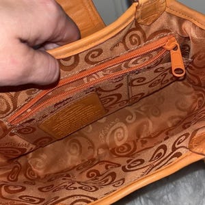 May include: Orange leather handbag with a patterned interior lining. The bag features a zippered pocket and a small leather tag. The zipper and stitching are in a matching orange color. The bag is open, revealing the interior.