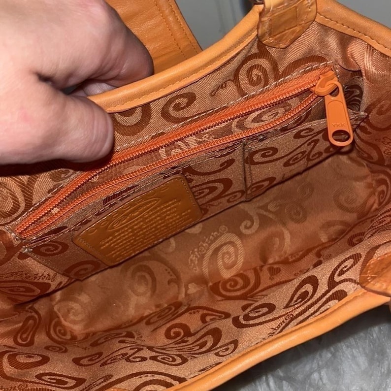 May include: Orange leather handbag with a patterned interior lining. The bag features a zippered pocket and a small leather tag. The zipper and stitching are in a matching orange color. The bag is open, revealing the interior.