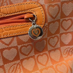 May include: Close-up of an orange handbag with a heart pattern. The bag features a zipper with a faux leather accent and a silver charm with a heart design. The bag's pattern includes orange hearts on a lighter orange background.