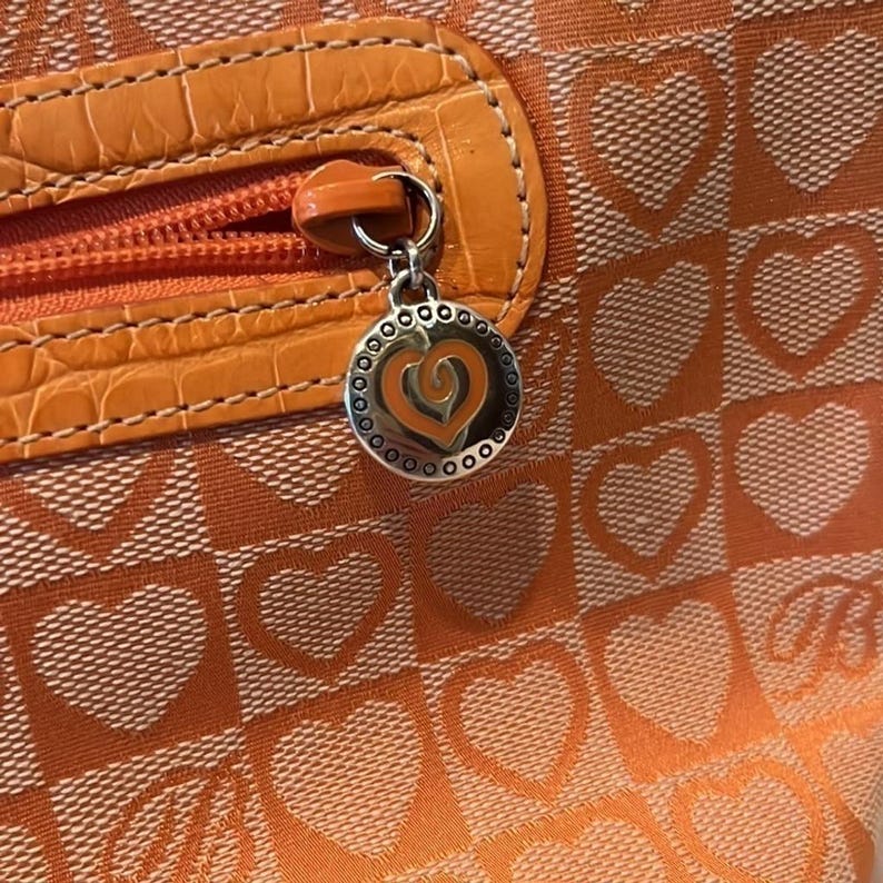 May include: Close-up of an orange handbag with a heart pattern. The bag features a zipper with a faux leather accent and a silver charm with a heart design. The bag's pattern includes orange hearts on a lighter orange background.