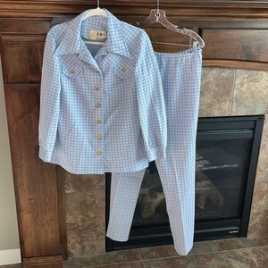 May include: A vintage two-piece outfit featuring a light blue and white plaid pattern. The set includes a long-sleeved button-down shirt with gold buttons and a pair of matching pants. The outfit is hanging on a hanger.