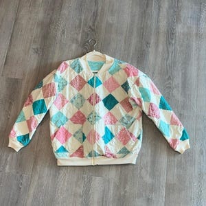 May include: A quilted bomber jacket with a cream-colored base and a diamond pattern in shades of pink, teal, and blue. The jacket has a zip-up front, ribbed cuffs, and a ribbed hem. The jacket is hanging on a white hanger.