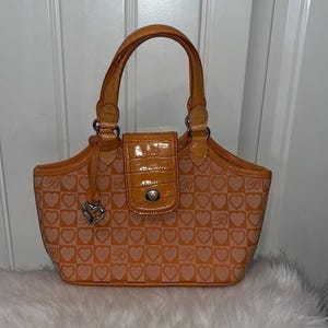 May include: Orange handbag with a heart pattern and a top handle. The bag features a flap closure and a silver heart charm. The bag's design includes a repeating heart and letter pattern.