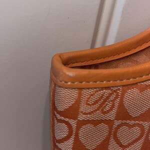 May include: Close-up of an orange and white patterned handbag. The bag features a repeating heart and letter "B" design. The trim is a solid orange color.
