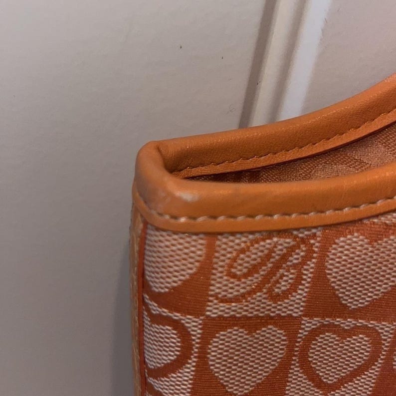 May include: Close-up of an orange and white patterned handbag. The bag features a repeating heart and letter "B" design. The trim is a solid orange color.