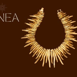 Könnte beinhalten: Eine goldfarbene Statement-Halskette mit einem einzigartigen, stacheligen Design. Die Halskette besteht aus zahlreichen, unregelmäßigen, länglichen Teilen, die nach außen strahlen. Das Wort "LINEA" wird in weißer Schrift auf der linken Seite mit einer sonnenartigen Grafik darüber angezeigt.