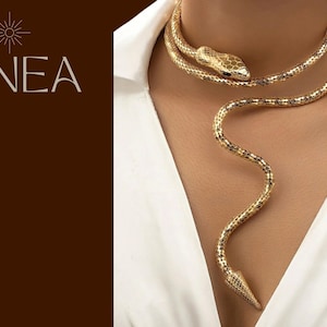 Puede incluir: Collar de serpiente dorado con un diseño detallado de cabeza y cola. El collar se envuelve alrededor del cuello y cae sobre el pecho. La palabra "LINEA" está impresa en blanco en el lado izquierdo de la imagen.