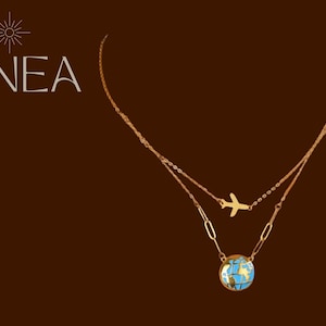 Puede incluir: Collar dorado en capas con un pequeño dije de avión y un colgante de globo terráqueo. El globo presenta detalles en azul y dorado. La palabra "LINEA" está en texto blanco en la esquina superior izquierda. El fondo es de color marrón oscuro.