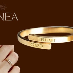 May include: A gold-coloured ring with the words "TRUST GOD" engraved on it. The ring has a wrap-around design. The word "LINEA" is in the upper left corner of the image.