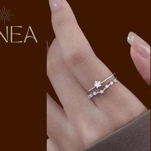 Könnte beinhalten: Nahaufnahme von zwei silbernen Ringen an einem Finger. Der obere Ring hat ein sternförmiges Design, während der untere Ring eine Reihe kleiner, funkelnder Steine aufweist. Das Wort "LINEA" ist oben links sichtbar.