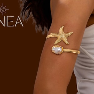 May include: Gold-toned arm cuff featuring a starfish and a pearl-like embellishment. The cuff is worn on the upper arm. The word "LINEA" is displayed in a stylized font at the top left corner of the image.