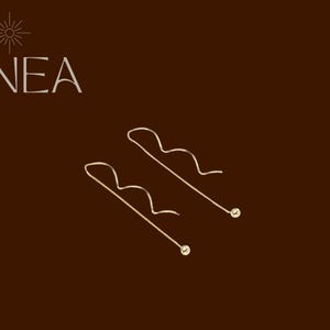 Puede incluir: Un par de pendientes colgantes dorados con un diseño ondulado, cada uno con una pequeña cuenta redonda al final. La palabra "LINEA" está impresa en blanco en la esquina superior izquierda sobre un fondo marrón.