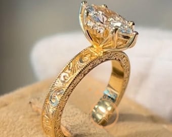Könnte beinhalten: Ein goldener Ring mit einem verzierten, gravierten Band und einem großen, birnenförmigen Diamantencluster. Der Ring ist auf einer beigefarbenen Oberfläche platziert, die seine komplizierten Details und funkelnden Edelsteine hervorhebt. Das Design des Rings ist klassisch und elegant.