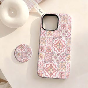 May include: A phone case and matching pop socket featuring a pink, yellow, and gray tile pattern. The case has a black camera cutout. The pop socket is round and has the same tile design. The items are on a white surface.