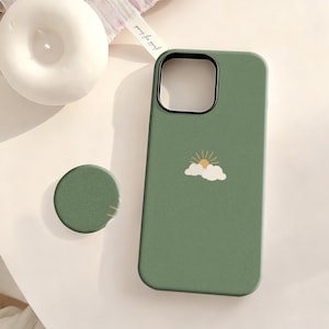 Puede incluir: Una funda de teléfono verde salvia con un agarre de teléfono redondo a juego. La funda presenta un pequeño gráfico de un sol y nubes. La funda del teléfono tiene un anillo de cámara negro.