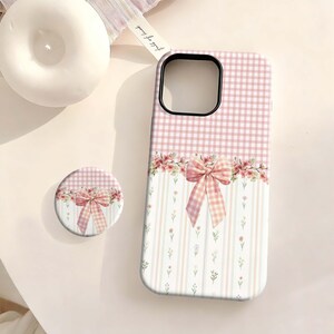 Pode incluir: Uma capa de telefone rosa e branca com um pop socket a condizer. A capa apresenta um padrão xadrez na parte superior e um design floral e de laço na parte inferior. O pop socket tem um design semelhante.