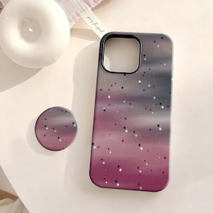 May include: A phone case and matching pop socket with a starry night design. The case and pop socket have a gradient of purple and gray with white and dark purple stars. The phone case has a black border.