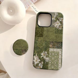 May include: A rectangular phone case and a circular phone grip featuring a patchwork design of various green ornate patterns and white lily floral illustrations. The phone case has a black camera cutout and the phone grip matches the case design.