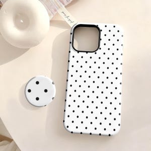 May include: White phone case and matching pop socket with a black polka dot pattern. The phone case has a black camera cutout. A white candle and a ribbon are in the background.