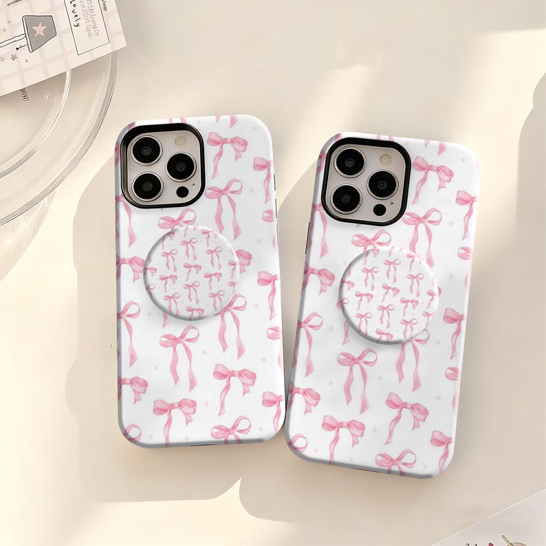May include: Two white phone cases with a pink bow pattern and matching pop sockets. The bows are watercolor style and scattered across the case. The pop sockets have a smaller bow pattern.