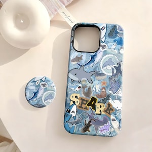 May include: A blue phone case and matching pop socket featuring various cartoon shark illustrations. The word "SHARK" is printed in a brown and yellow font on the phone case. The case and pop socket have a light blue background.