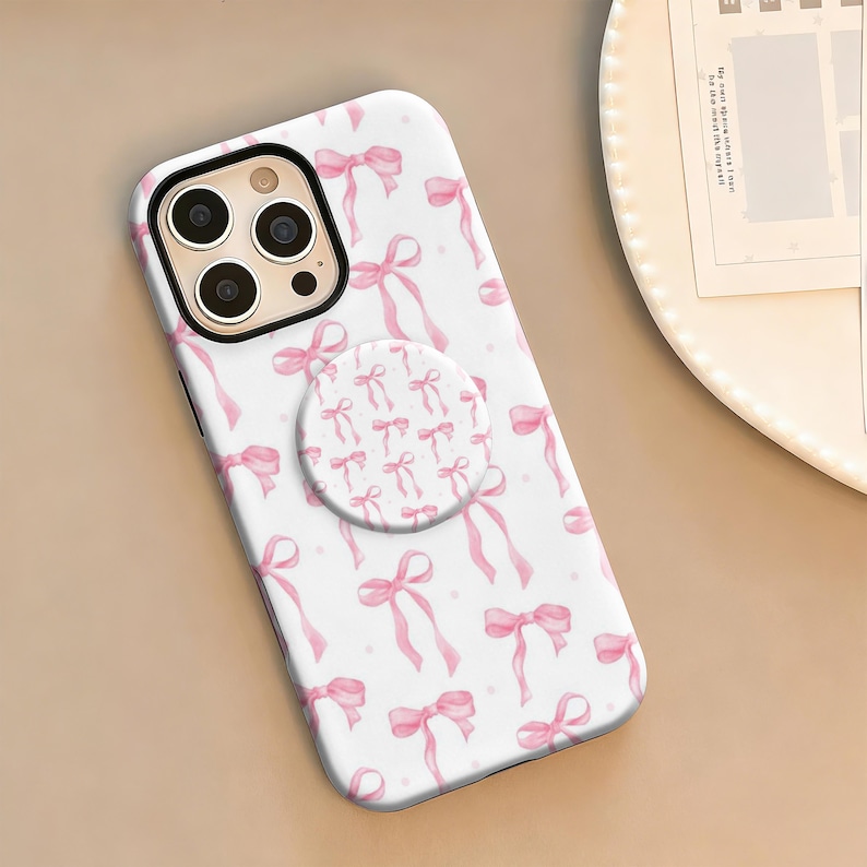 May include: White iPhone case with a pink bow pattern and a matching pop socket. The case features a repeating design of pink bows and ribbons on a white background. The pop socket has a smaller version of the bow pattern.