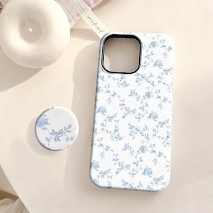 May include: A white phone case and matching phone grip featuring a delicate blue floral pattern. The case has a black camera cutout and is next to a white candle. The phone grip is round and has the same floral design.