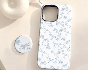 May include: A white phone case and matching phone grip featuring a delicate blue floral pattern. The case has a black camera cutout and is next to a white candle. The phone grip is round and has the same floral design.