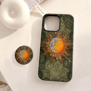Può includere: Una custodia rettangolare per telefono e un supporto circolare con sfondo verde scuro e motivi a vortice. Il design presenta un'illustrazione centrale di sole e luna con un centro a spirale arancione brillante, raggi dorati e una luna crescente blu.