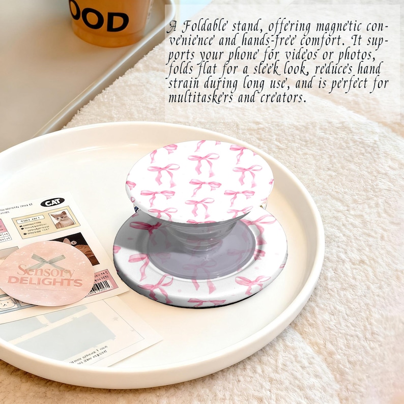 May include: A white and pink phone stand with a bow pattern. The stand is on a white plate with other items, including a sticker that says "Sensory Delights". The stand folds flat for easy storage.