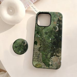 Puede incluir: Funda de teléfono verde y agarre a juego con un diseño celestial de mariposas, lunas y estrellas. La funda tiene un borde verde oscuro y un recorte para la cámara. El agarre redondo tiene un diseño similar.