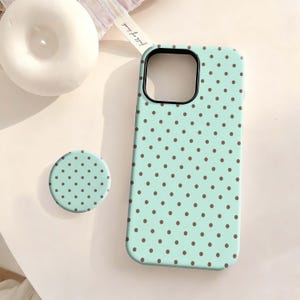 May include: A mint green phone case and matching pop socket with a repeating pattern of small brown polka dots. The phone case has a black camera lens border. The pop socket is round and has the same polka dot design.