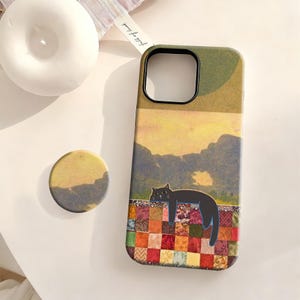 May include: A rectangular phone case and a circular phone grip featuring a black cat resting on a multicolored patchwork quilt. The background design displays a yellow sky above dark green mountains. The phone case has a black camera cutout.