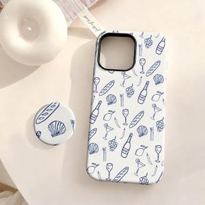 May include: White phone case and matching pop socket with a blue line art pattern of wine bottles, grapes, bread, and other food items. The phone case has a black border. A white candle and a ribbon are in the background.