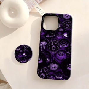 May include: A purple and black phone case and matching pop socket featuring an eye pattern. The phone case has a camera cutout and is made of a hard plastic material. The pop socket is round and has a similar design.
