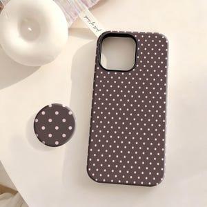 Könnte beinhalten: Eine dunkelbraune Handyhülle und ein passender Pop-Socket, beide mit einem sich wiederholenden Muster aus kleinen, hellrosa Punkten. Die Handyhülle hat einen schwarzen Kameraausschnitt. Der Pop-Socket ist rund und hat das gleiche Punktmuster.