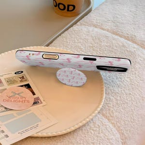 May include: White phone case and matching pop socket with a pink bow pattern. The phone case is on a white plate with a small piece of paper that says "Sensory Delights".